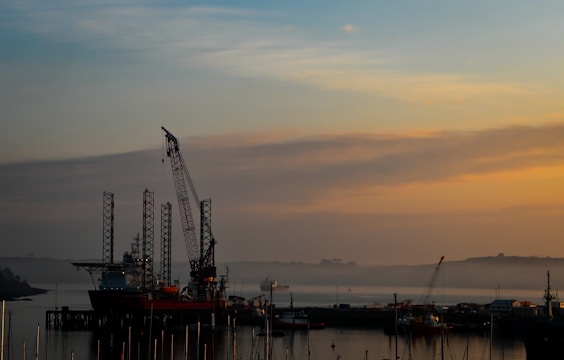 A peaceful harbor scene at sunset with an offshore rig silhouetted against a colorful sky. Cranes and the rig’s tall structures are prominent against the soft oranges and blues of the sunset. Calm water reflects the muted colors of the sky, and faint outlines of distant boats and land are visible.