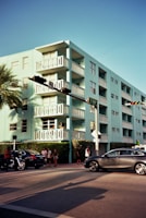 A pastel blue apartment building with multiple balconies stands prominently on a street corner. Several cars and a motorcycle are visible on the street, while a small group of pedestrians gathers on the sidewalk. A palm tree and some traffic lights add to the urban scene.