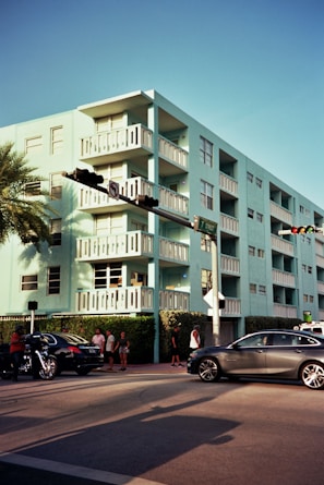 A pastel blue apartment building with multiple balconies stands prominently on a street corner. Several cars and a motorcycle are visible on the street, while a small group of pedestrians gathers on the sidewalk. A palm tree and some traffic lights add to the urban scene.