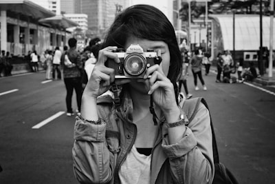 A person holding a camera up to their face, standing in the middle of a street surrounded by a crowd of people. The image is in black and white, enhancing the urban atmosphere.