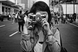 A person holding a camera up to their face, standing in the middle of a street surrounded by a crowd of people. The image is in black and white, enhancing the urban atmosphere.