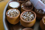 Small clay pots filled with various grains and seeds are arranged on a tray. Some pots contain popped sorghum, while others hold different grains and nuts. A rolled-up paper or cloth is tucked to the side.