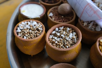 Small clay pots filled with various grains and seeds are arranged on a tray. Some pots contain popped sorghum, while others hold different grains and nuts. A rolled-up paper or cloth is tucked to the side.