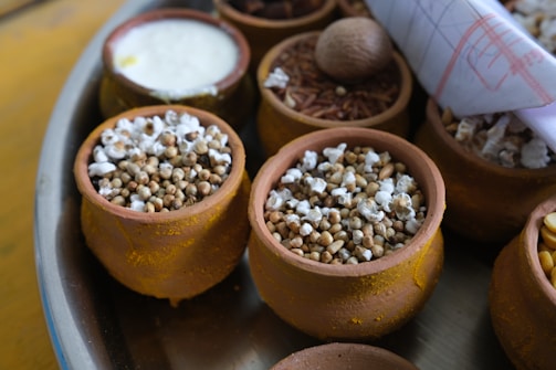Small clay pots filled with various grains and seeds are arranged on a tray. Some pots contain popped sorghum, while others hold different grains and nuts. A rolled-up paper or cloth is tucked to the side.