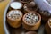 Small clay pots filled with various grains and seeds are arranged on a tray. Some pots contain popped sorghum, while others hold different grains and nuts. A rolled-up paper or cloth is tucked to the side.