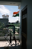 An Indian flag is waving outside on a balcony, with a view of a residential area and some greenery in the background. A clothes drying rack with various clothes is positioned near the railing. The weather appears clear with a blue sky and scattered clouds.