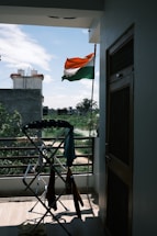 An Indian flag is waving outside on a balcony, with a view of a residential area and some greenery in the background. A clothes drying rack with various clothes is positioned near the railing. The weather appears clear with a blue sky and scattered clouds.