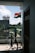 An Indian flag is waving outside on a balcony, with a view of a residential area and some greenery in the background. A clothes drying rack with various clothes is positioned near the railing. The weather appears clear with a blue sky and scattered clouds.