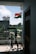 An Indian flag is waving outside on a balcony, with a view of a residential area and some greenery in the background. A clothes drying rack with various clothes is positioned near the railing. The weather appears clear with a blue sky and scattered clouds.