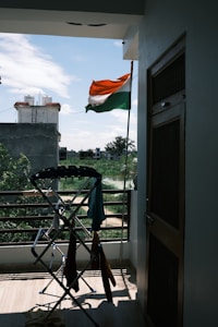 An Indian flag is waving outside on a balcony, with a view of a residential area and some greenery in the background. A clothes drying rack with various clothes is positioned near the railing. The weather appears clear with a blue sky and scattered clouds.