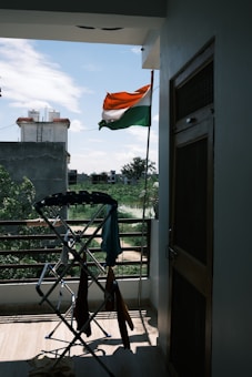 An Indian flag is waving outside on a balcony, with a view of a residential area and some greenery in the background. A clothes drying rack with various clothes is positioned near the railing. The weather appears clear with a blue sky and scattered clouds.