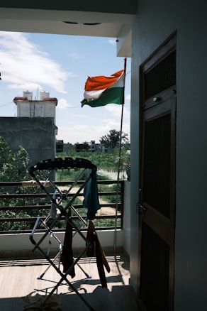 An Indian flag is waving outside on a balcony, with a view of a residential area and some greenery in the background. A clothes drying rack with various clothes is positioned near the railing. The weather appears clear with a blue sky and scattered clouds.