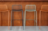 Four colorful metal bar stools with minimalist design are arranged in front of a brown, wooden wall. The stools are colored in alternating shades of orange and teal. The setting appears to be modern and industrial, likely a part of a bar or a cafe.