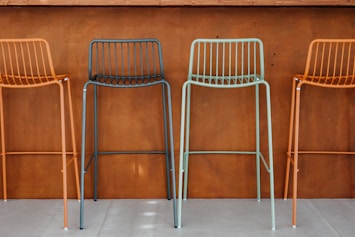 Four colorful metal bar stools with minimalist design are arranged in front of a brown, wooden wall. The stools are colored in alternating shades of orange and teal. The setting appears to be modern and industrial, likely a part of a bar or a cafe.