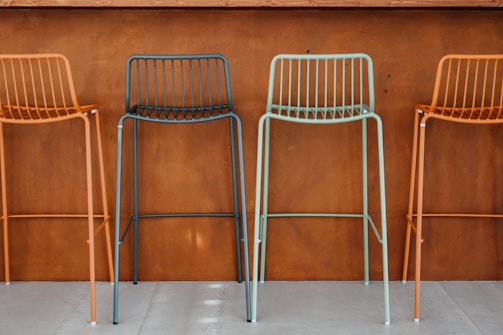 Four colorful metal bar stools with minimalist design are arranged in front of a brown, wooden wall. The stools are colored in alternating shades of orange and teal. The setting appears to be modern and industrial, likely a part of a bar or a cafe.