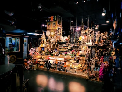A vibrant shop filled with a variety of decorative items, including clocks, statues, paintings, and trinkets. The display is densely packed, creating a rich, eclectic atmosphere. There are hanging ornaments, shelves crowded with memorabilia, and various colorful objects scattered throughout the scene. The setting is dimly lit, adding an intriguing and mysterious ambiance.
