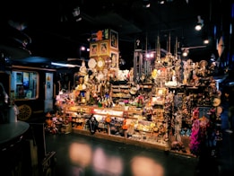 A vibrant shop filled with a variety of decorative items, including clocks, statues, paintings, and trinkets. The display is densely packed, creating a rich, eclectic atmosphere. There are hanging ornaments, shelves crowded with memorabilia, and various colorful objects scattered throughout the scene. The setting is dimly lit, adding an intriguing and mysterious ambiance.