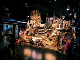 A vibrant shop filled with a variety of decorative items, including clocks, statues, paintings, and trinkets. The display is densely packed, creating a rich, eclectic atmosphere. There are hanging ornaments, shelves crowded with memorabilia, and various colorful objects scattered throughout the scene. The setting is dimly lit, adding an intriguing and mysterious ambiance.