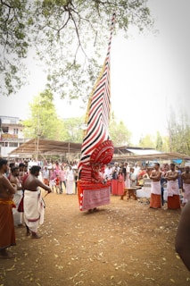 A traditional cultural performance featuring an elaborate costume with a tall, colorful headpiece and patterned red and white designs. The performer is surrounded by a group of spectators and participants, some wearing traditional attire, indicating a festive or ceremonial setting. The environment is outdoors with trees and a natural ground covering.