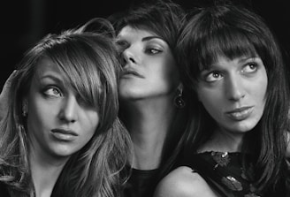 Three women are captured in a black and white photograph. Each has distinct hairstyles and facial expressions, creating a sense of individuality and character. The lighting emphasizes their features and expressions, giving a dramatic and artistic feel.
