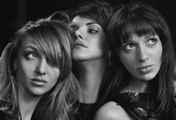 Three women are captured in a black and white photograph. Each has distinct hairstyles and facial expressions, creating a sense of individuality and character. The lighting emphasizes their features and expressions, giving a dramatic and artistic feel.