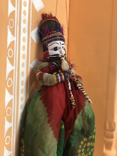 A vibrant traditional puppet with a painted face, wearing an elaborate headdress and colorful clothing. It is suspended against a decorated wall with geometric patterns in white on an earthy background.