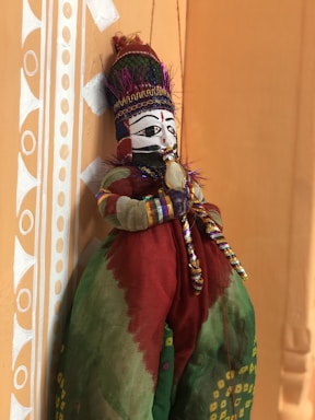 A vibrant traditional puppet with a painted face, wearing an elaborate headdress and colorful clothing. It is suspended against a decorated wall with geometric patterns in white on an earthy background.