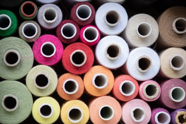 Various colorful spools of thread arranged closely together, showcasing a wide range of colors including green, pink, red, orange, and white. The spools are viewed from above, emphasizing the circular shapes of the thread wrapped around them.