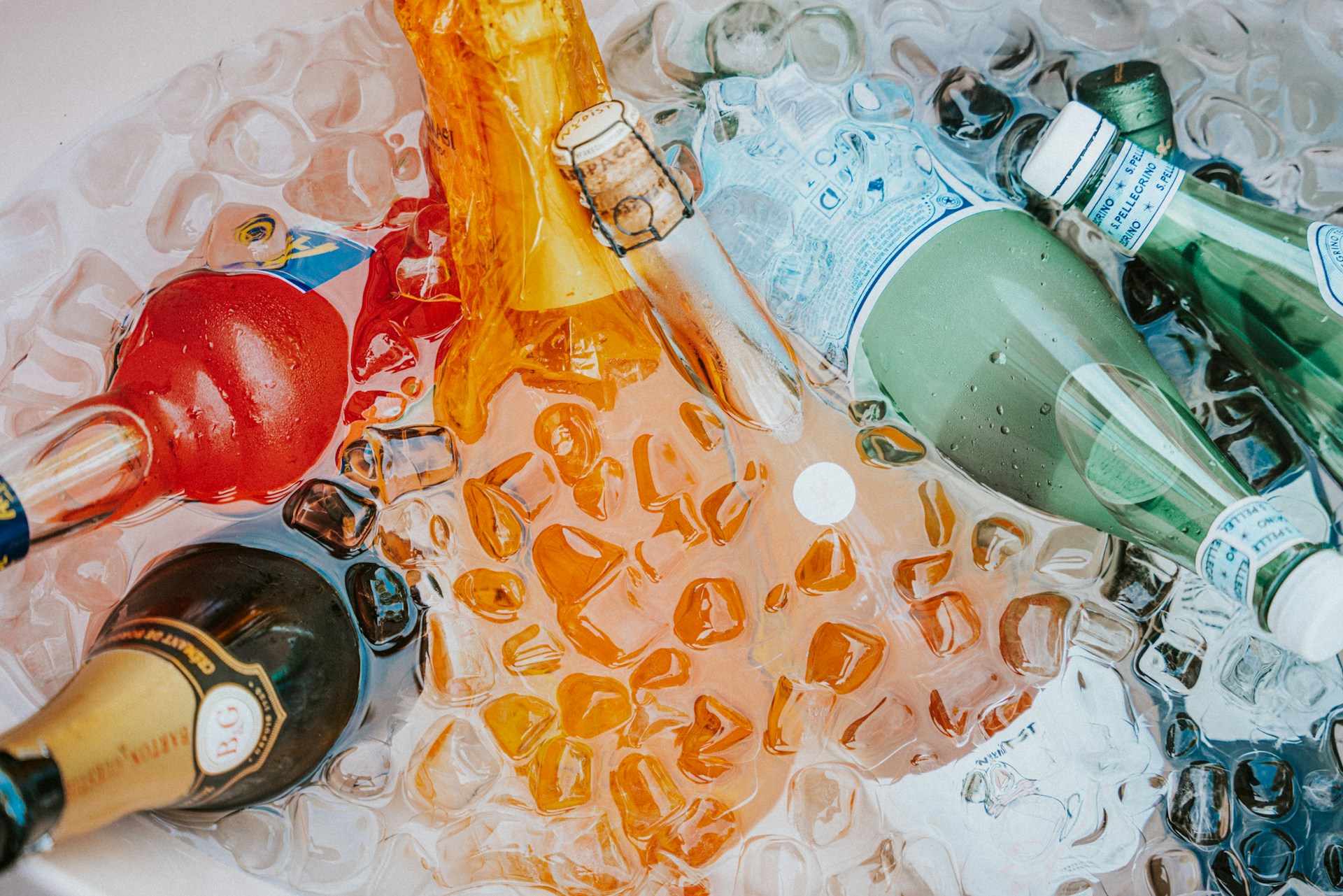 Several bottles, including champagne and sparkling water, are chilled in a bin filled with ice cubes. The bottles are of various colors and shapes, partially submerged in melting ice, creating a refreshing and festive scene.