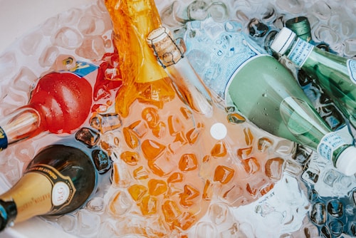 Several bottles, including champagne and sparkling water, are chilled in a bin filled with ice cubes. The bottles are of various colors and shapes, partially submerged in melting ice, creating a refreshing and festive scene.