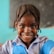 A young child with braided hair is smiling warmly. The child is wearing a checkered blue and white shirt with a blue apron over it. The background is blurred, featuring a teal hue and a soft blackboard.