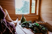 A cozy sauna interior with wooden paneling and a bench. A person is resting in the sauna with a relaxed posture, legs extended. Near them, there are branches of green leaves and a woolen hat with text on it. A window offers a view of the outside greenery, allowing natural light to illuminate the space. A basket containing stones is also visible.