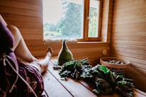 A cozy sauna interior with wooden paneling and a bench. A person is resting in the sauna with a relaxed posture, legs extended. Near them, there are branches of green leaves and a woolen hat with text on it. A window offers a view of the outside greenery, allowing natural light to illuminate the space. A basket containing stones is also visible.