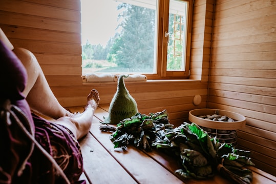 A cozy sauna interior with wooden paneling and a bench. A person is resting in the sauna with a relaxed posture, legs extended. Near them, there are branches of green leaves and a woolen hat with text on it. A window offers a view of the outside greenery, allowing natural light to illuminate the space. A basket containing stones is also visible.