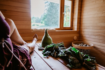 A cozy sauna interior with wooden paneling and a bench. A person is resting in the sauna with a relaxed posture, legs extended. Near them, there are branches of green leaves and a woolen hat with text on it. A window offers a view of the outside greenery, allowing natural light to illuminate the space. A basket containing stones is also visible.