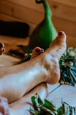 A pair of feet with painted toenails resting on a wooden floor surrounded by green leaves and a large green gourd in the background.
