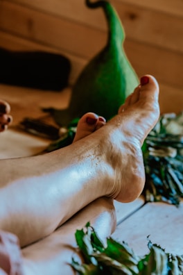 A pair of feet with painted toenails resting on a wooden floor surrounded by green leaves and a large green gourd in the background.