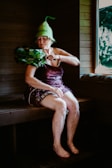 An elderly woman is sitting in a wooden sauna, wearing a unique green hat and utilizing a bundle of leafy branches known as a birch whisk. She appears to be in a relaxed state, with her eyes closed. A window reveals a view of the greenery outside.