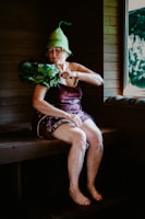An elderly woman is sitting in a wooden sauna, wearing a unique green hat and utilizing a bundle of leafy branches known as a birch whisk. She appears to be in a relaxed state, with her eyes closed. A window reveals a view of the greenery outside.