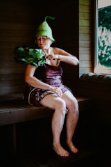 An elderly woman is sitting in a wooden sauna, wearing a unique green hat and utilizing a bundle of leafy branches known as a birch whisk. She appears to be in a relaxed state, with her eyes closed. A window reveals a view of the greenery outside.