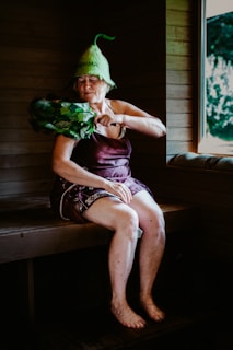 An elderly woman is sitting in a wooden sauna, wearing a unique green hat and utilizing a bundle of leafy branches known as a birch whisk. She appears to be in a relaxed state, with her eyes closed. A window reveals a view of the greenery outside.
