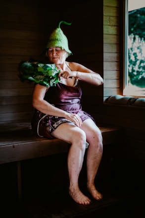 An elderly woman is sitting in a wooden sauna, wearing a unique green hat and utilizing a bundle of leafy branches known as a birch whisk. She appears to be in a relaxed state, with her eyes closed. A window reveals a view of the greenery outside.