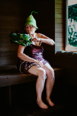 An elderly woman is sitting in a wooden sauna, wearing a unique green hat and utilizing a bundle of leafy branches known as a birch whisk. She appears to be in a relaxed state, with her eyes closed. A window reveals a view of the greenery outside.