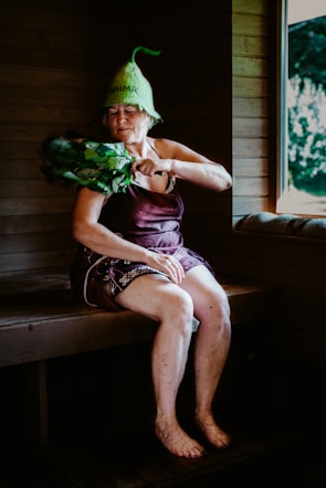 An elderly woman is sitting in a wooden sauna, wearing a unique green hat and utilizing a bundle of leafy branches known as a birch whisk. She appears to be in a relaxed state, with her eyes closed. A window reveals a view of the greenery outside.