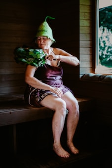 An elderly woman is sitting in a wooden sauna, wearing a unique green hat and utilizing a bundle of leafy branches known as a birch whisk. She appears to be in a relaxed state, with her eyes closed. A window reveals a view of the greenery outside.