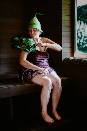 An elderly woman is sitting in a wooden sauna, wearing a unique green hat and utilizing a bundle of leafy branches known as a birch whisk. She appears to be in a relaxed state, with her eyes closed. A window reveals a view of the greenery outside.