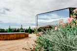 A serene backyard setting featuring a wooden deck with a hot tub, surrounded by green shrubs and pink flowers. There is a large reflective mirror capturing the blue sky and fluffy clouds. A row of tall trees lines the edge of the property, adding a touch of privacy and nature.