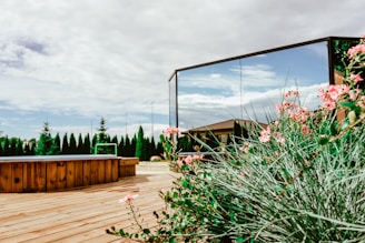 A serene backyard setting featuring a wooden deck with a hot tub, surrounded by green shrubs and pink flowers. There is a large reflective mirror capturing the blue sky and fluffy clouds. A row of tall trees lines the edge of the property, adding a touch of privacy and nature.