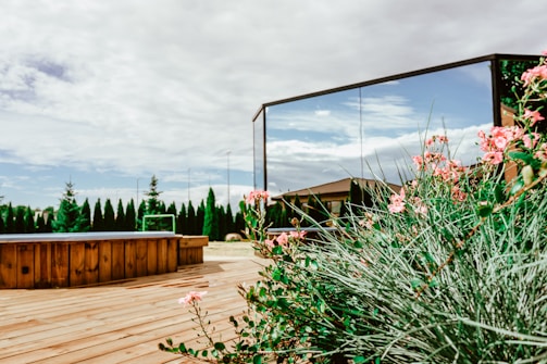 A serene backyard setting featuring a wooden deck with a hot tub, surrounded by green shrubs and pink flowers. There is a large reflective mirror capturing the blue sky and fluffy clouds. A row of tall trees lines the edge of the property, adding a touch of privacy and nature.