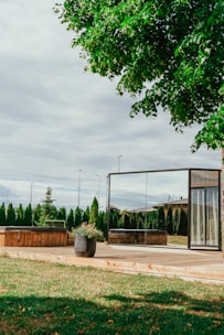 A serene outdoor scene featuring a wooden deck with a modern, glass-walled structure, nearby potted plants, and well-maintained green grass. Tall evergreen shrubs and trees create a natural boundary in the background, under a sky filled with light, fluffy clouds.